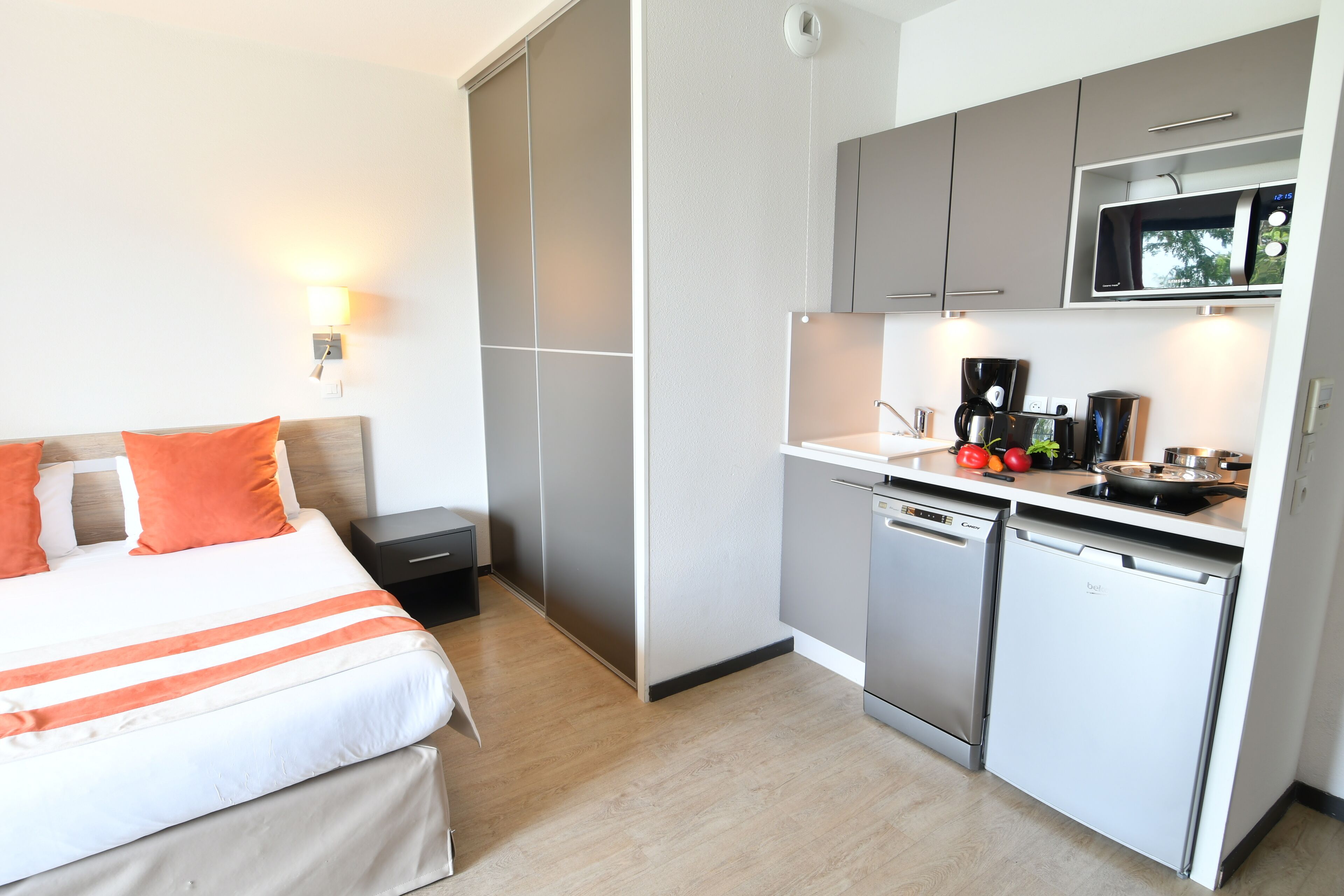Quarto de hotel com cama, mesa de cabeceira, escrivaninha e cozinha compacta equipada com micro-ondas, cafeteira e lava-louças.
