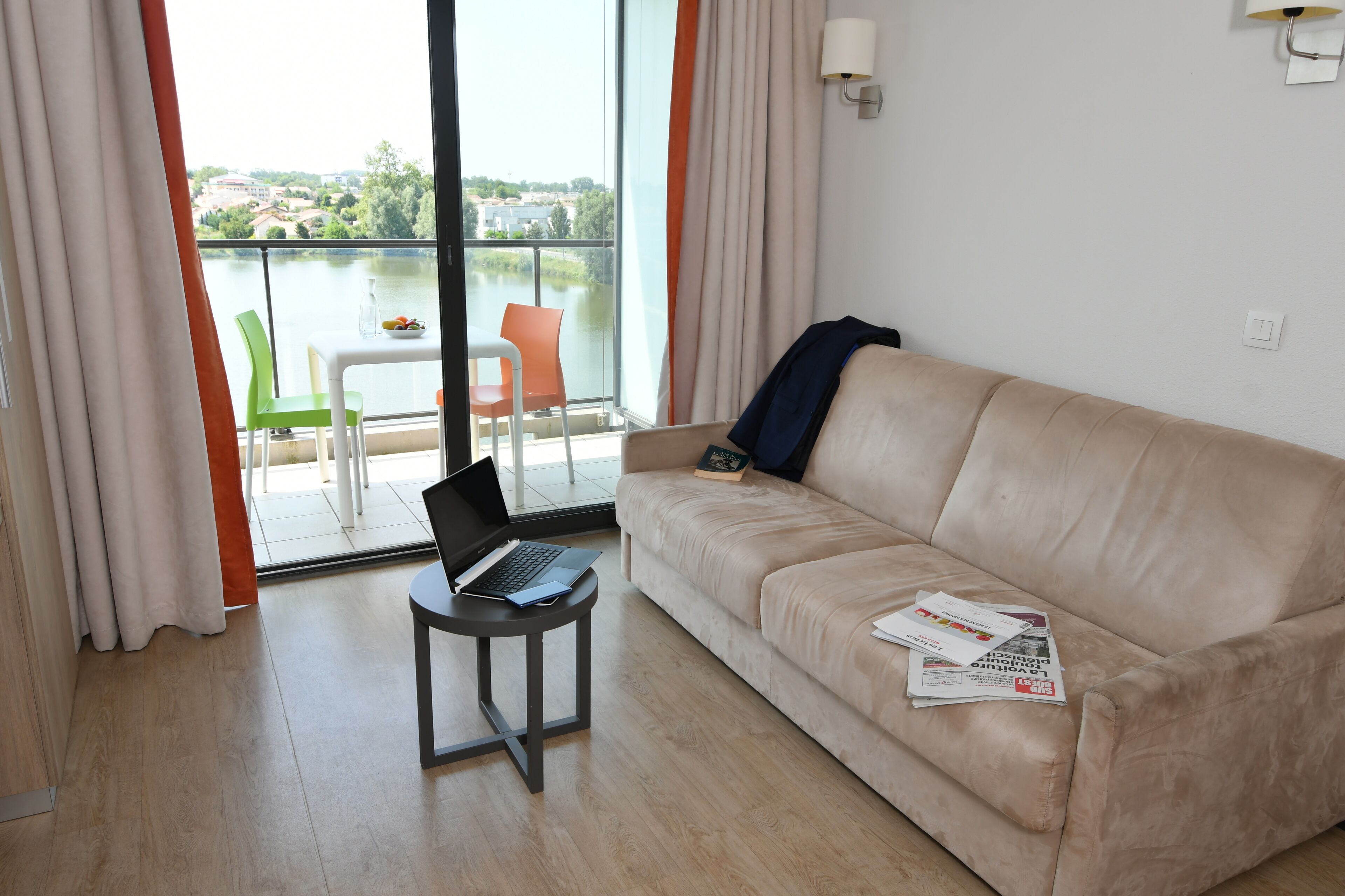 A room with a beige sofa, a small table with a laptop, and a view of a body of water through a large window.