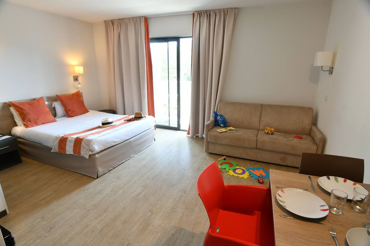 A hotel room with a bed, a sofa, a dining table with chairs, a lamp, and a window with curtains.