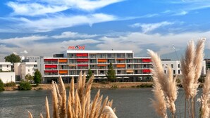 A multi-story hotel with balconies, a river in the foreground, and a clear sky. - Appart Hôtel Mer & Golf City Bordeaux Bruges (Bruges)