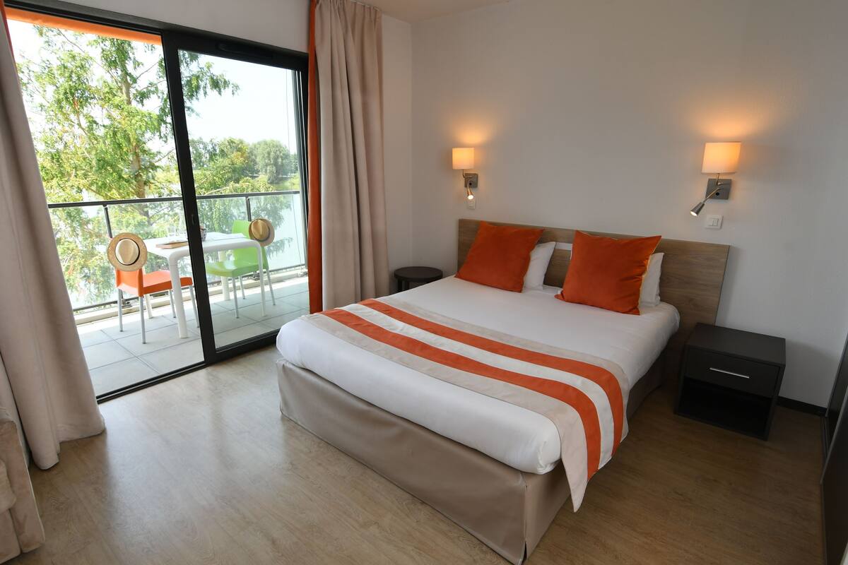 A modern hotel room with a large bed, two bedside tables, a balcony with a table and chairs, and a sliding glass door.