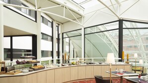Un hall d’hôtel moderne doté d’un plafond en verre, d’un espace buffet proposant divers plats et d’un coin salon avec des tables et des chaises.