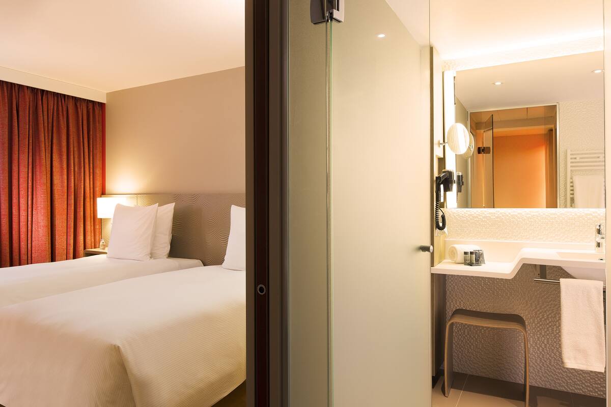 Habitación de hotel con dos camas, baño con lavamanos y espejo, y ventana con cortinas.