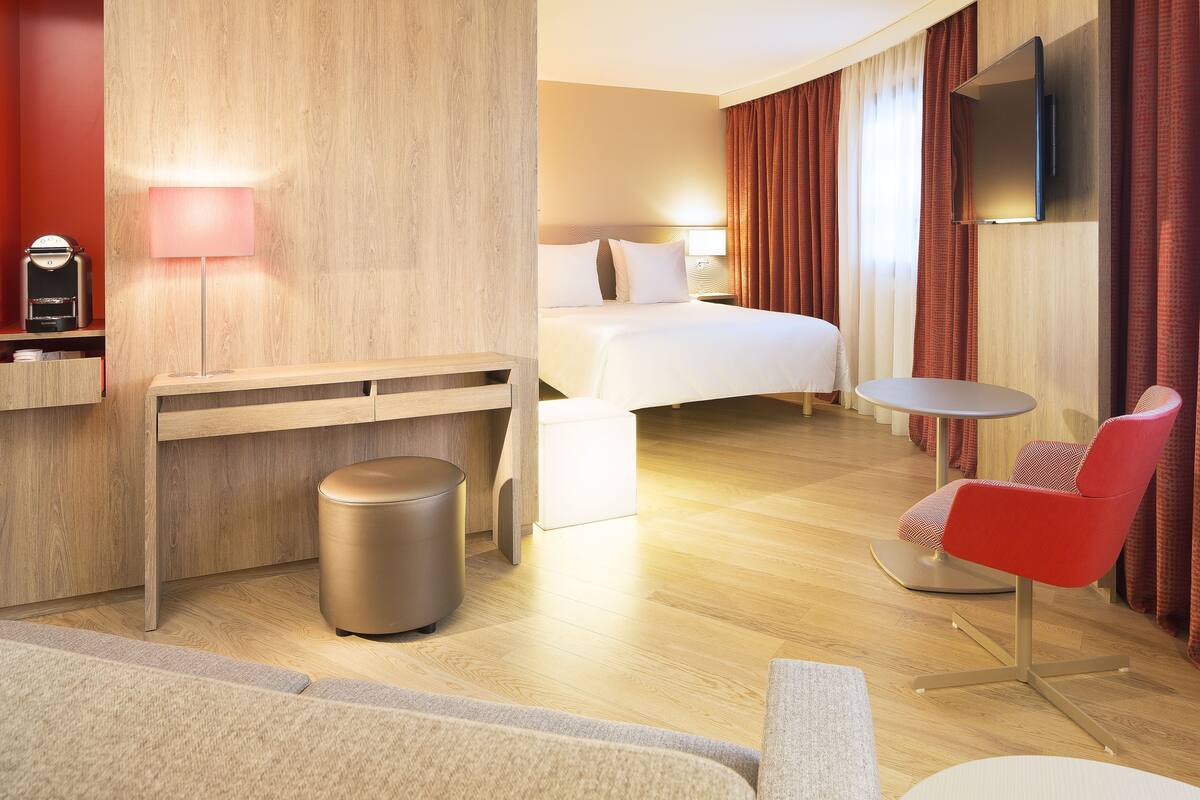 Habitación de hotel con cama, escritorio con lámpara, mesa redonda, una silla roja y televisor montado en la pared.