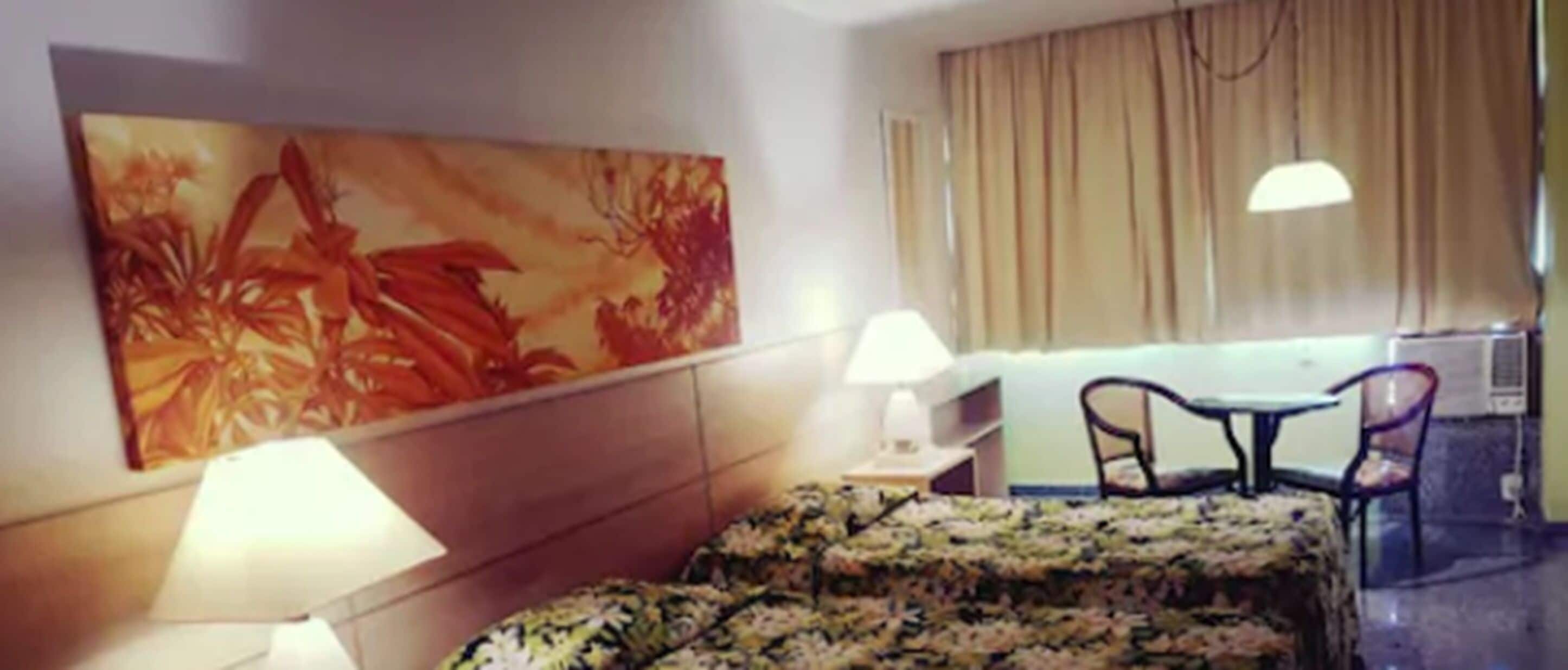 A hotel room with a bed, a desk, a chair, a lamp, and a large painting on the wall.