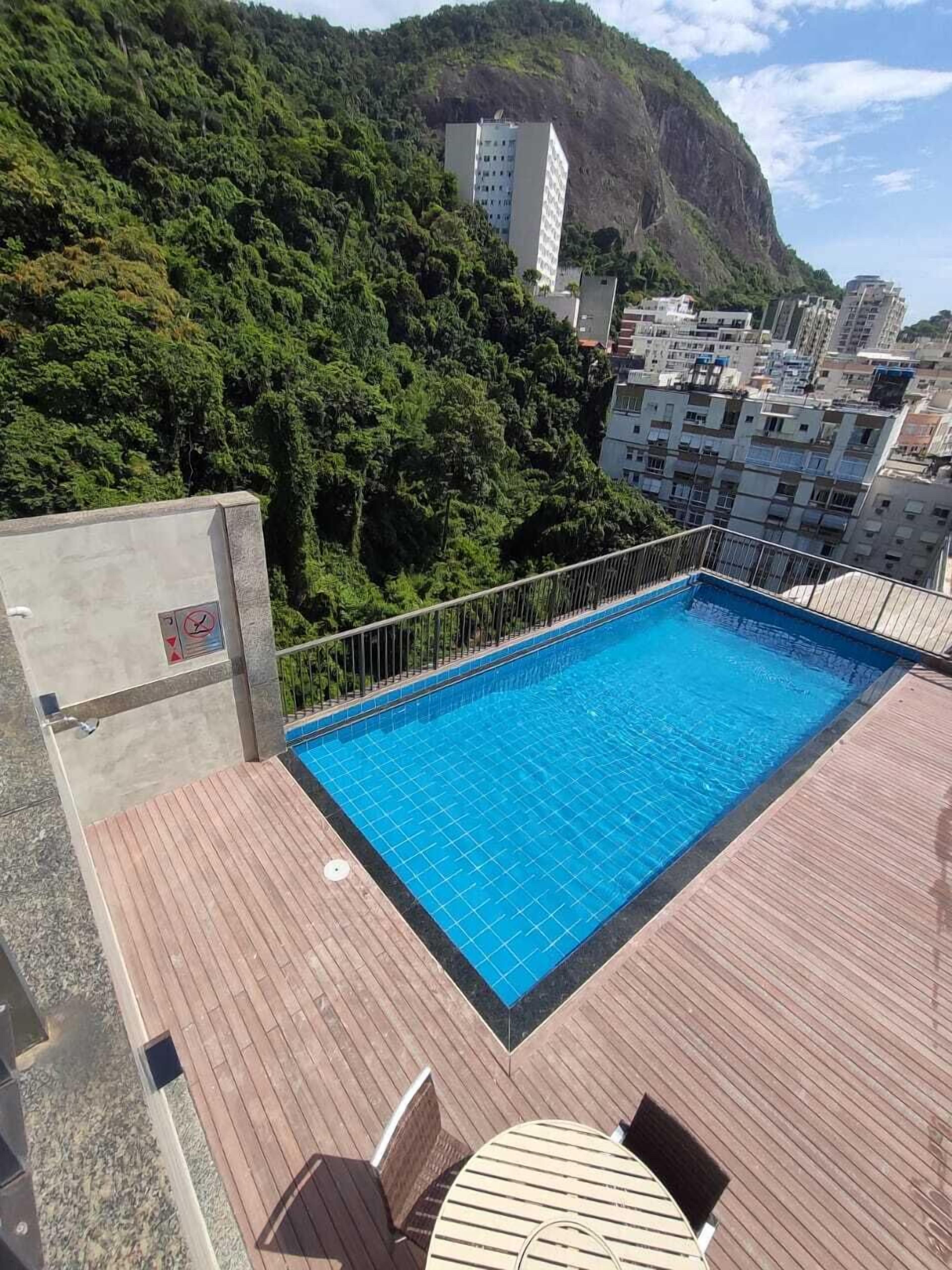 A rooftop pool with a wooden deck, surrounded by lush greenery and buildings.