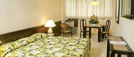 Habitació d'hotel amb llit, escriptori, cadira, làmpada i finestra amb cortines.