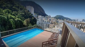 Ein Pool auf dem Dach mit einer Holzterrasse und Liegestühlen, umgeben von Gebäuden und einem Berg im Hintergrund.