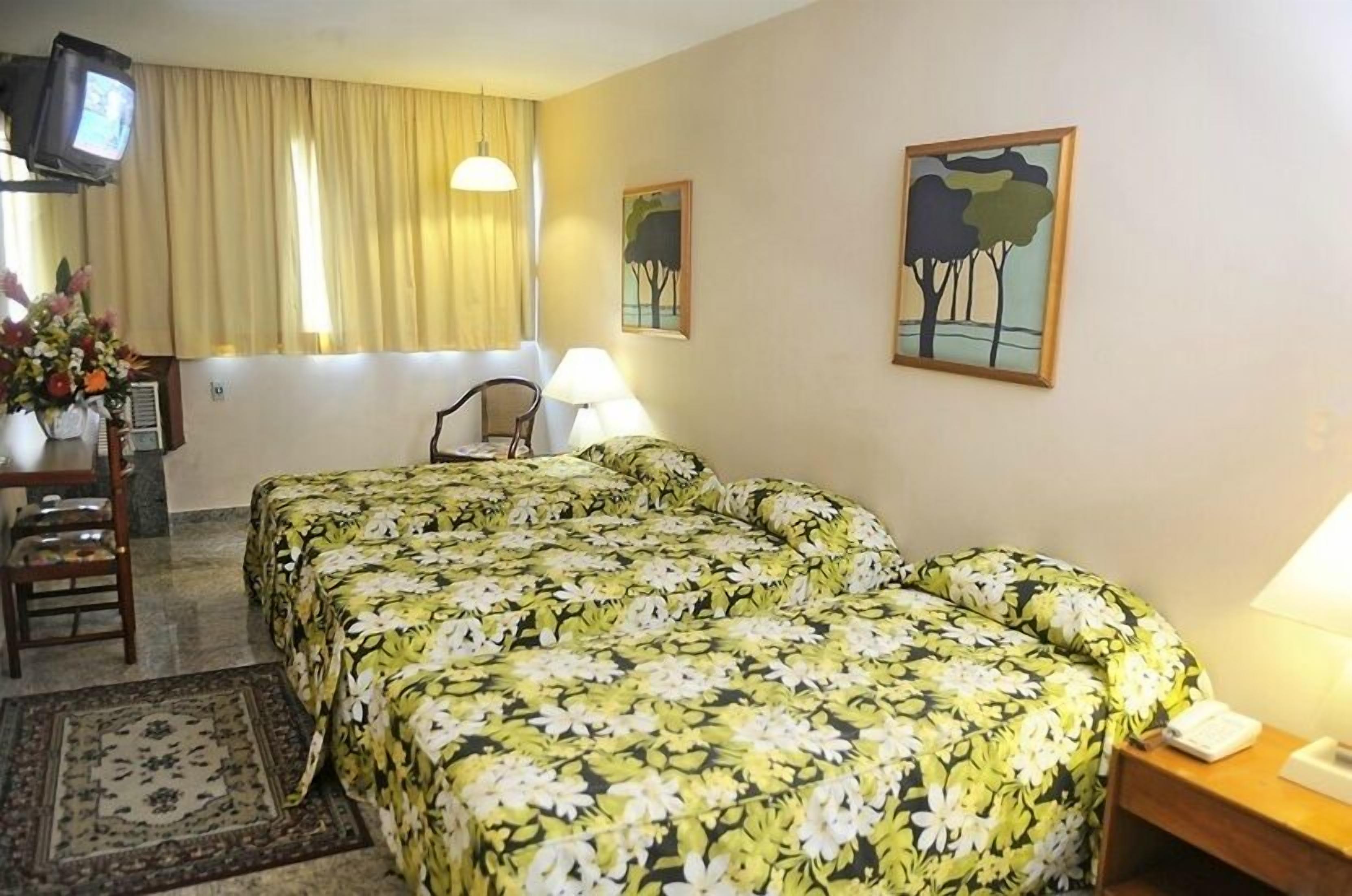 A hotel room with two beds, a desk with a chair, a TV, and two paintings on the wall.