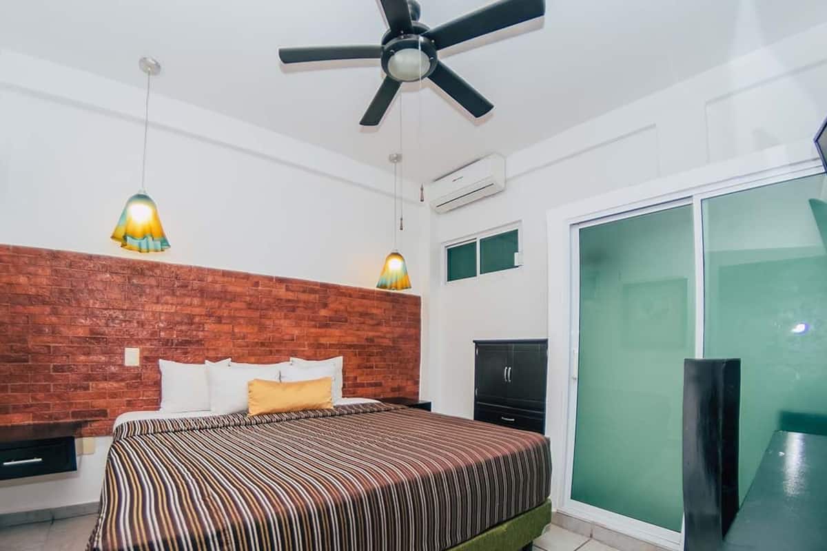 Ein Schlafzimmer mit einer Wand aus Ziegelsteinen, einem Bett mit gestreifter Bettwäsche, zwei Pendelleuchten, einem Deckenventilator, einem Fernseher und einer Schiebetür aus Glas.
