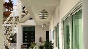 Een smalle, betegelde gang met witte muren en een trap aan de linkerkant. Er staan potplanten langs de gang en er hangt een lamp erboven.