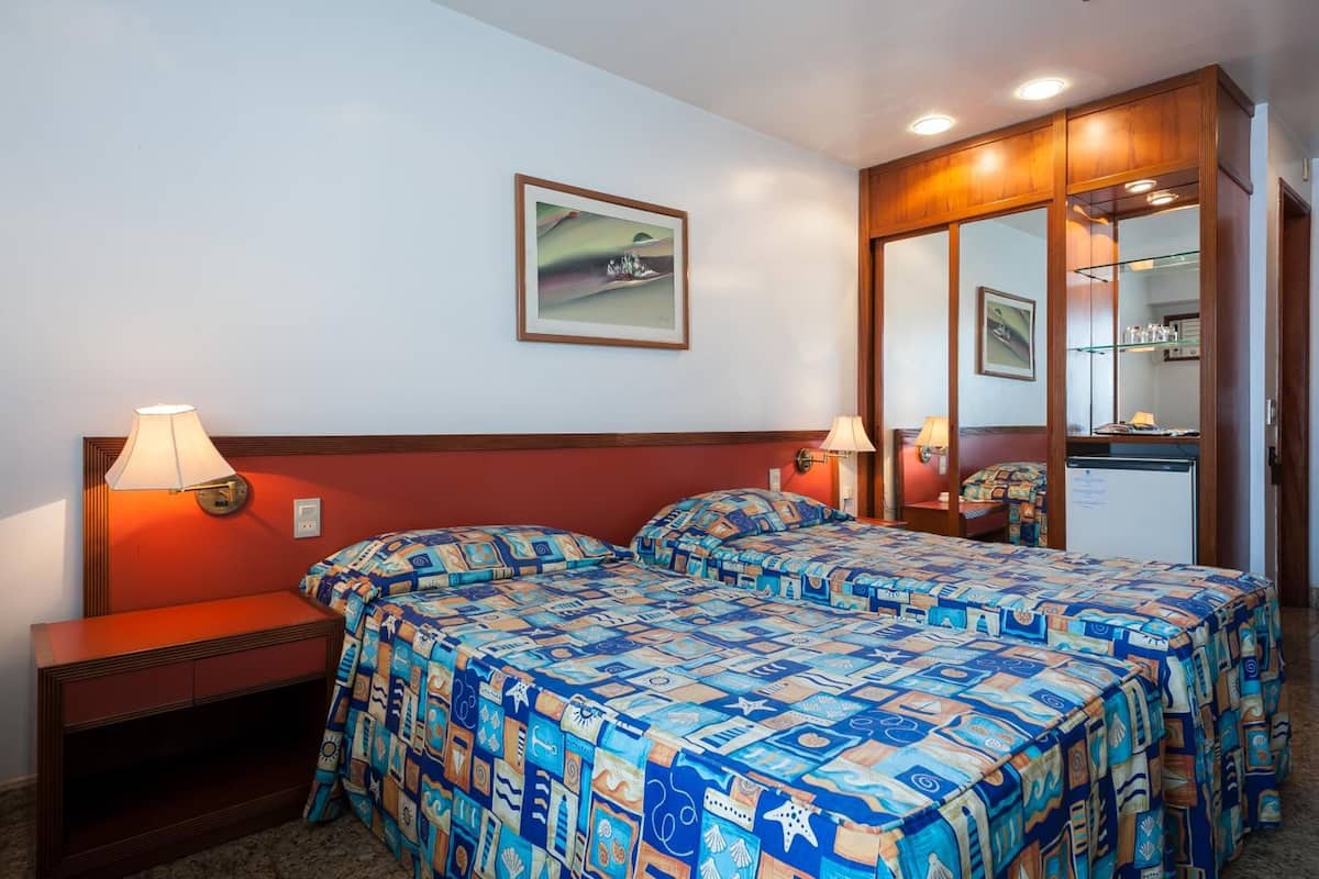 Une chambre d’hôtel avec deux lits, une table de chevet, une lampe, un miroir et un petit réfrigérateur.