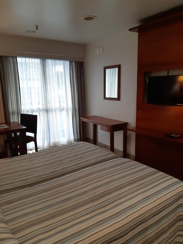 A hotel room with a large bed, a desk, a chair, a TV, and a window with curtains. - Oceano Copacabana Hotel (Rio de Janeiro)