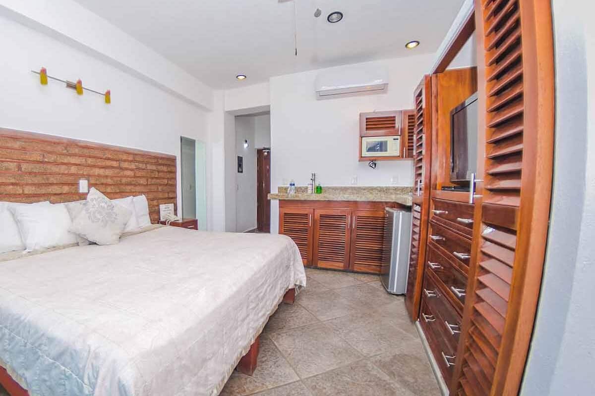 A bedroom with a large bed, wooden headboard, a small kitchenette with a microwave, and a bathroom visible in the background.