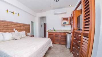 Un dormitorio con una cama grande, cabecera de madera, una pequeña kitchenette con microondas y un baño a simple vista.