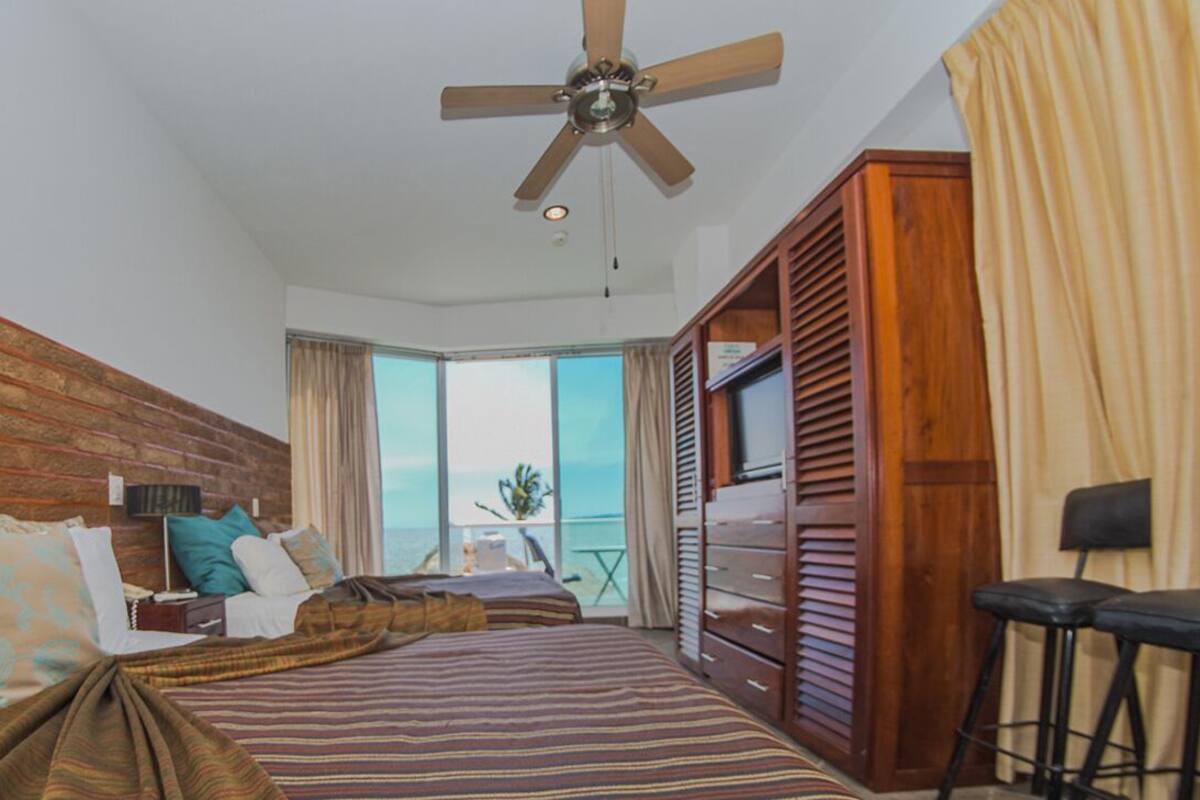 A hotel room with a large bed, a ceiling fan, a wooden headboard, a small table with a lamp, and a view of the outdoors.