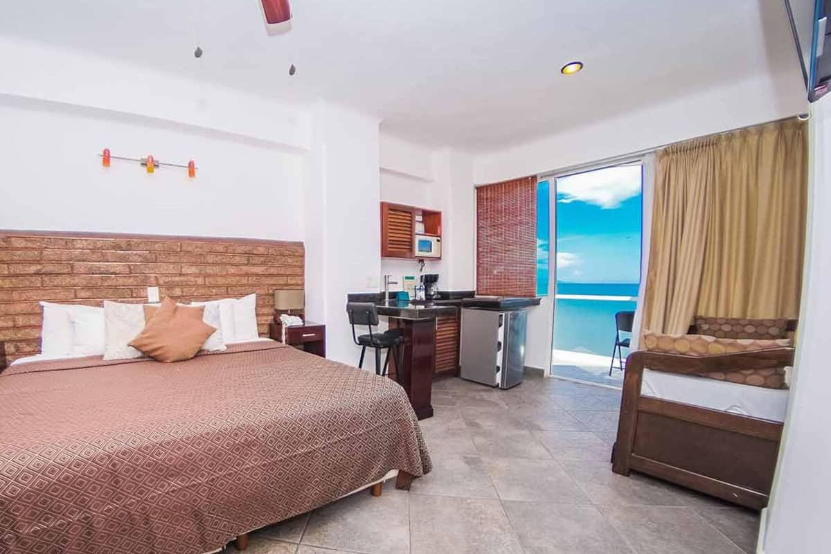 A hotel room with a large bed, a kitchenette, a balcony with a view, and a small dining area.