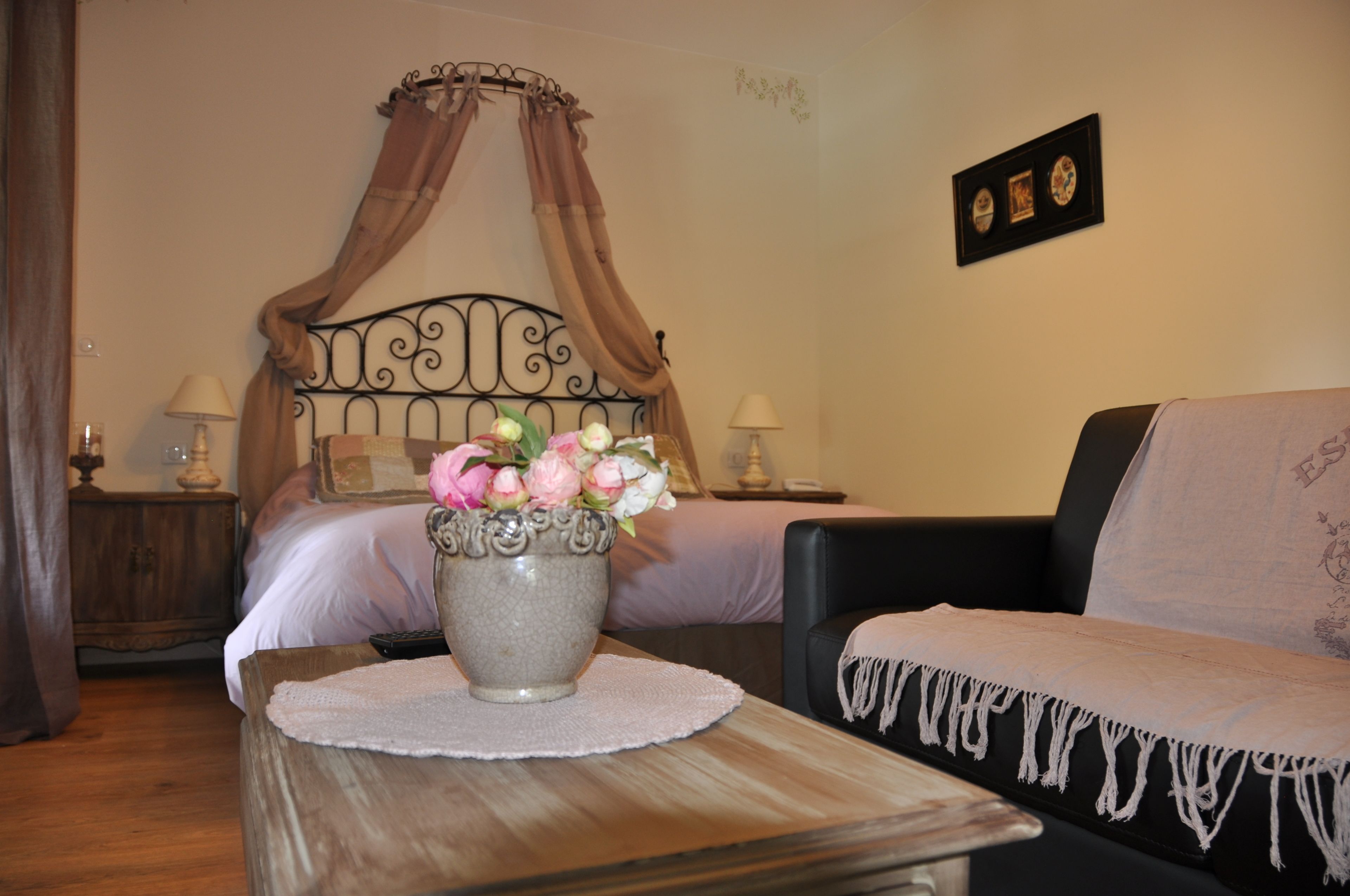 Un dormitorio con una cama con dosel, una mesita de noche con lámpara, un jarrón con flores y un cuadro en la pared.