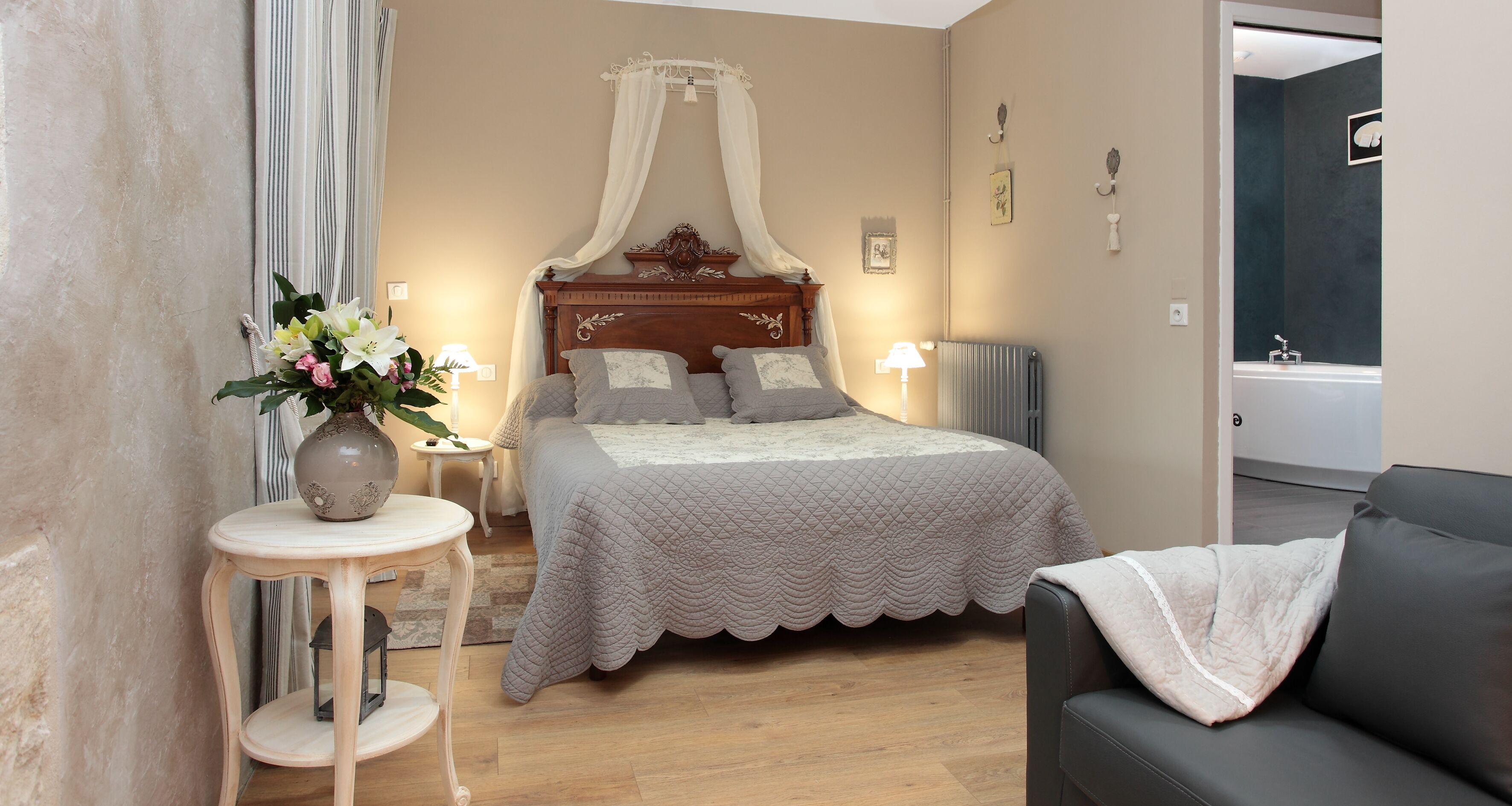Een slaapkamer met een hemelbed, een nachtkastje met een lamp, een bloevas en een grijze bank.