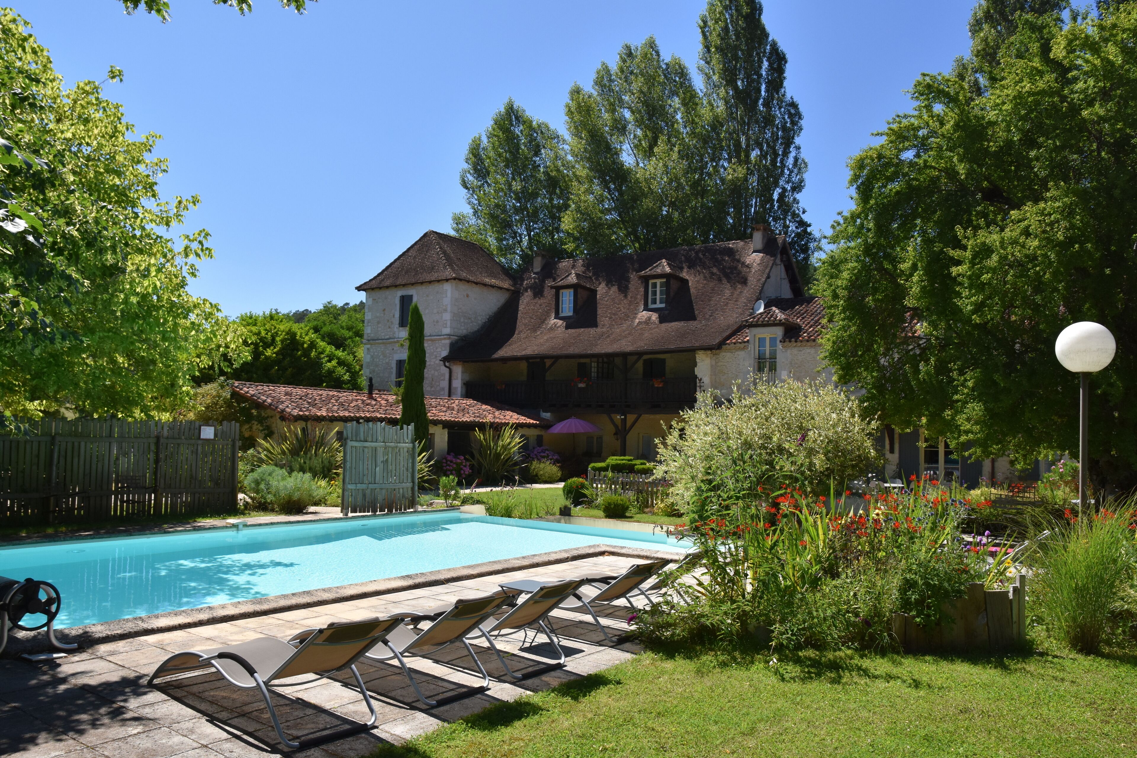 A swimming pool with lounge chairs, a wooden fence, and a large house with a tiled roof.