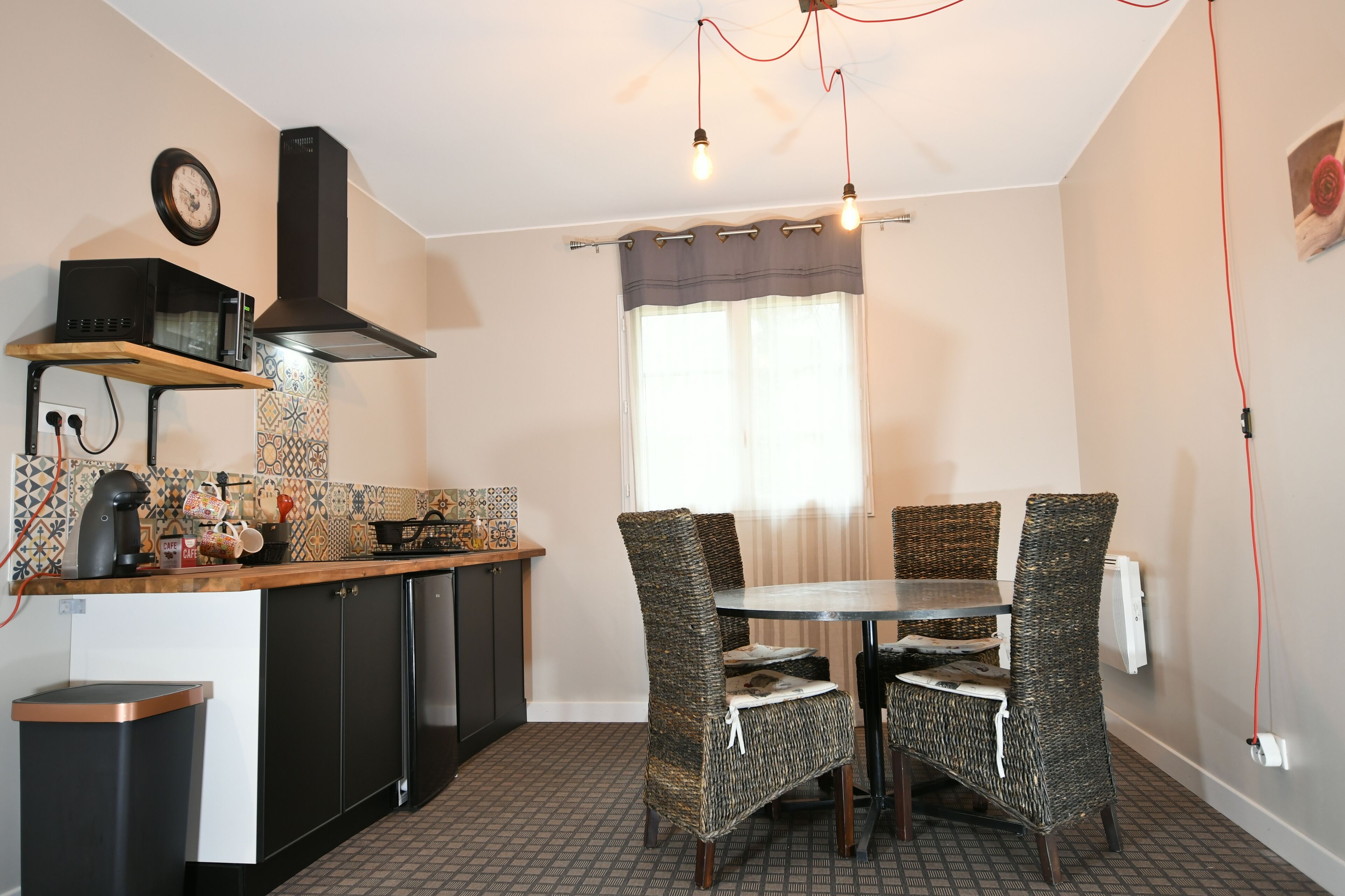 A compact kitchen with a microwave, stove, and sink. There is a dining table with wicker chairs and a window with curtains.
