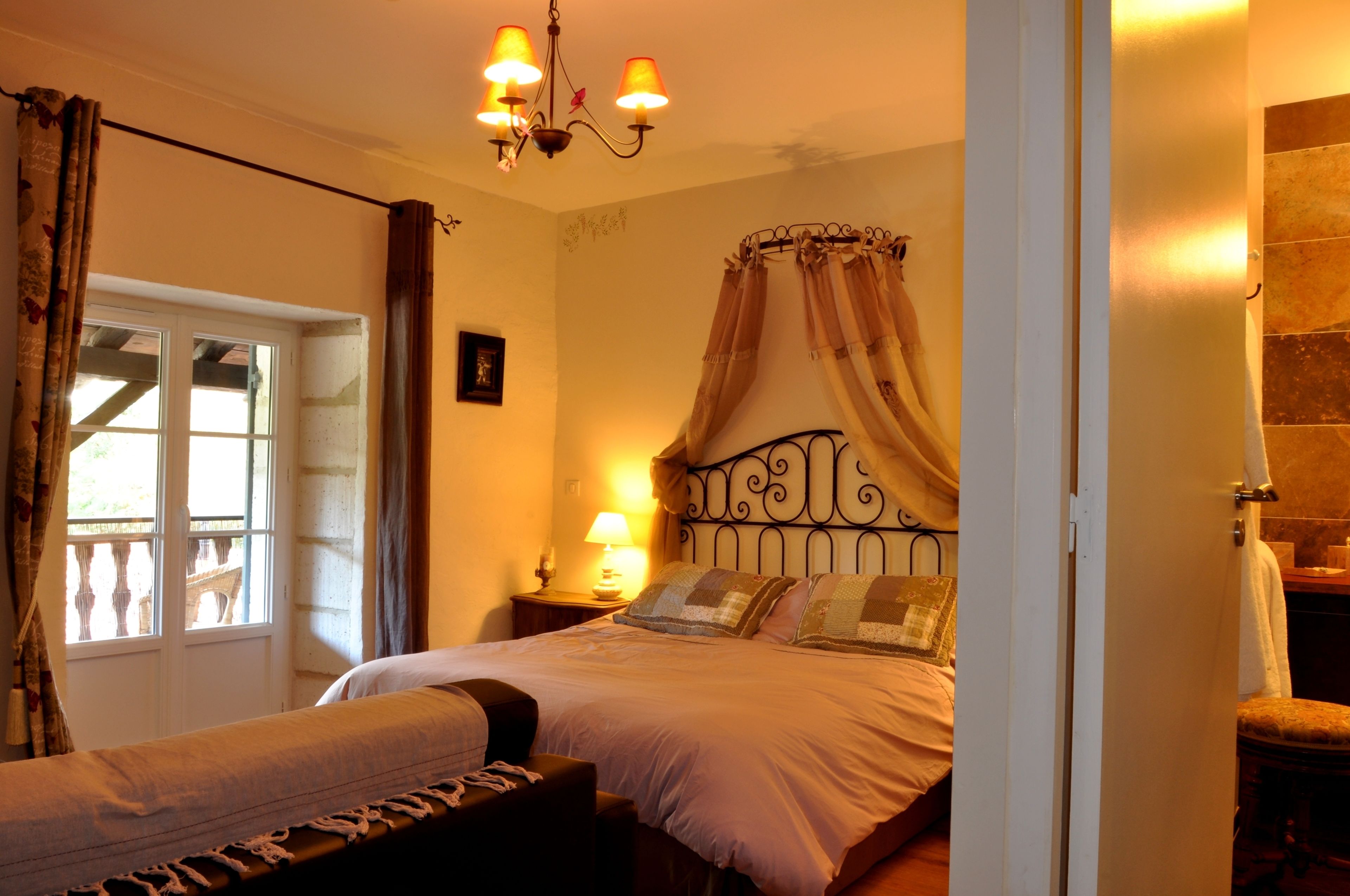 Un dormitorio con cama, mesita de noche con lámpara, ventana con cortinas y candelabro.