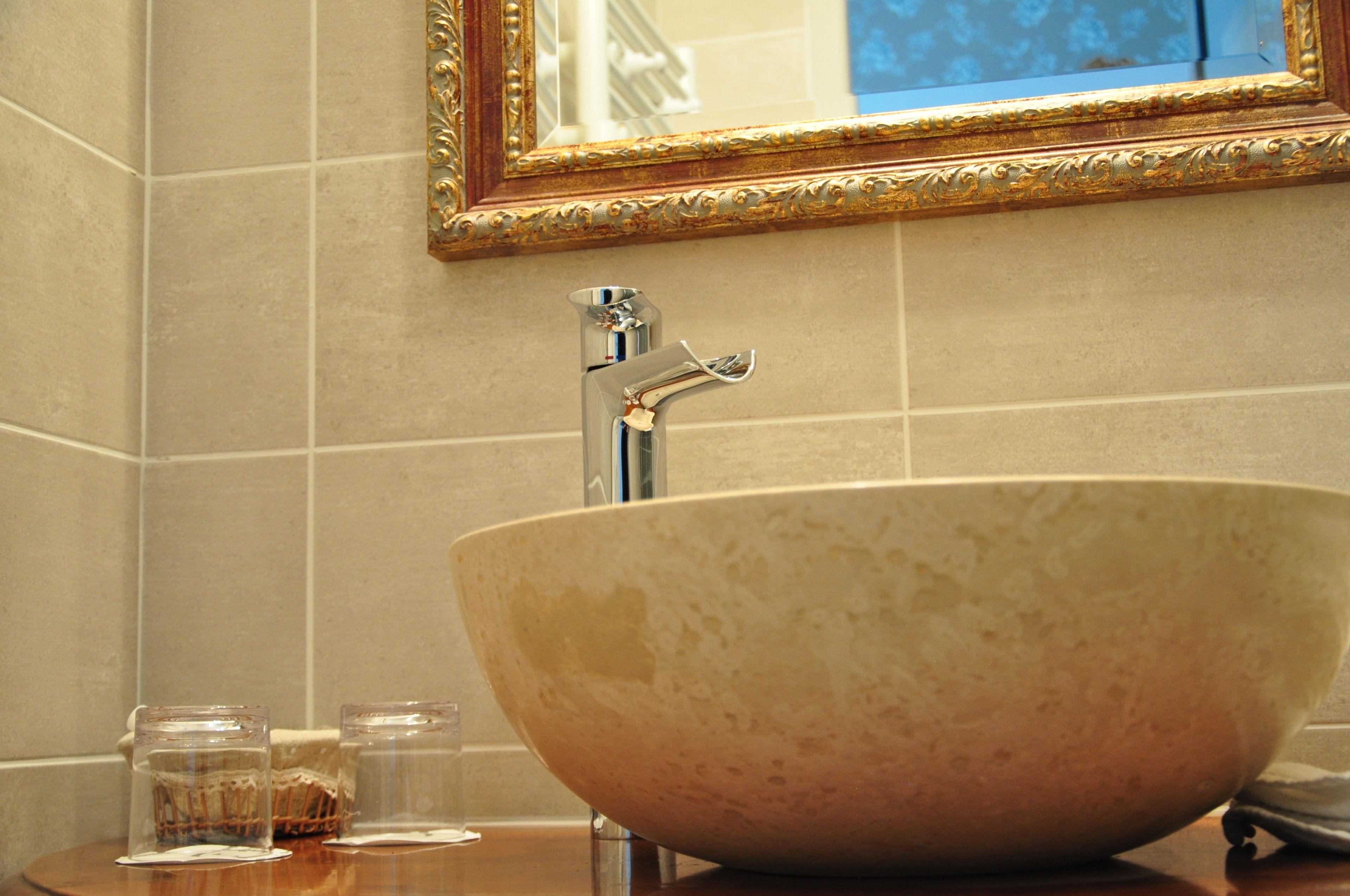 A bathroom with a large beige ceramic sink, a gold-framed mirror, and two clear glass bottles on the counter.