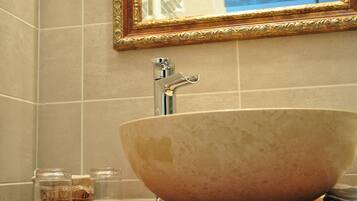 Un baño con un lavamanos grande de cerámica color beige, un espejo con marco dorado y dos botellas de vidrio transparente sobre la encimera.