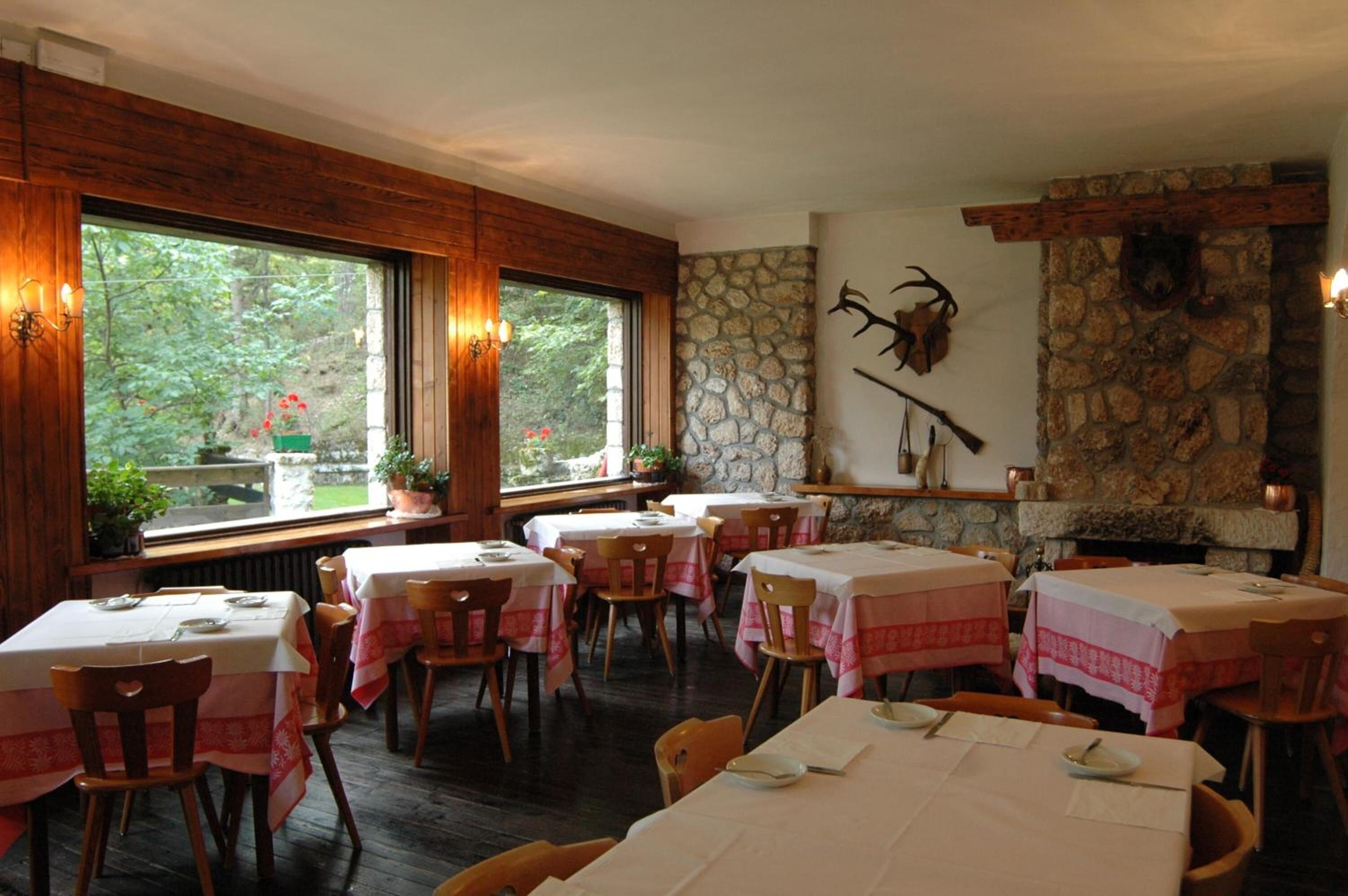 Un restaurant aux murs en bois, doté d’une cheminée en pierre et de grandes fenêtres donnant sur un espace verdoyant. Les tables sont dressées avec des nappes blanches et des serviettes rouges.