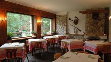 Un restaurant aux murs en bois, doté d’une cheminée en pierre et de grandes fenêtres donnant sur un espace verdoyant. Les tables sont dressées avec des nappes blanches et des serviettes rouges.