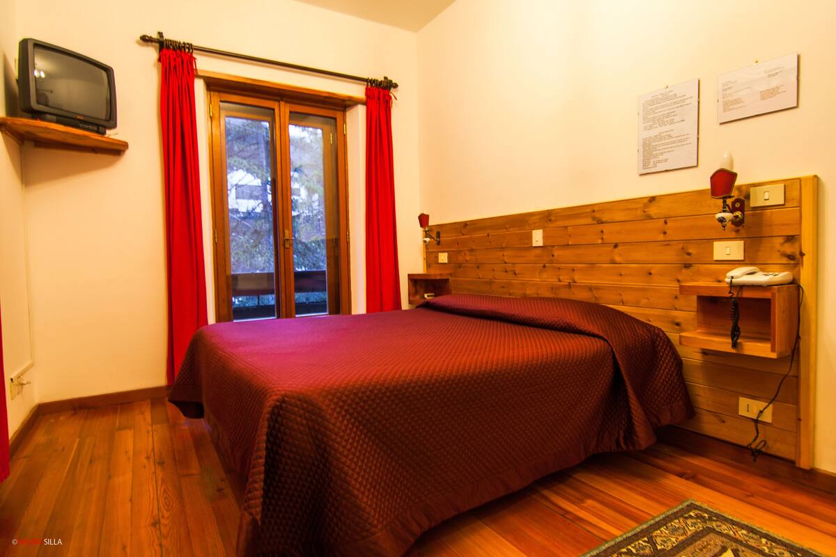A bedroom with a wooden headboard, a bed with a red blanket, a television on a shelf, and a window with red curtains.