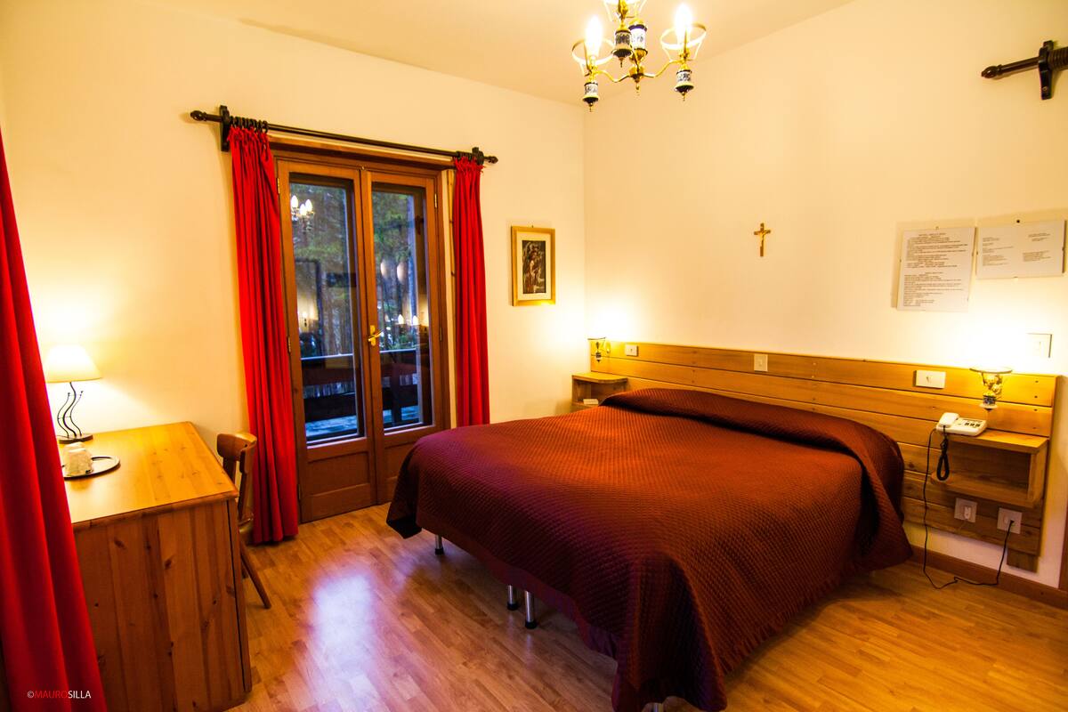 A bedroom with a wooden bed, a desk, a chair, a window with red curtains, a chandelier, and a cross on the wall.