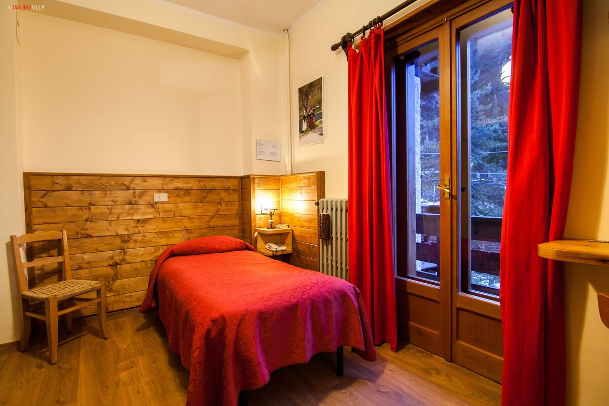 A room with a wooden headboard bed, a nightstand with a lamp, a chair, and a window with red curtains.