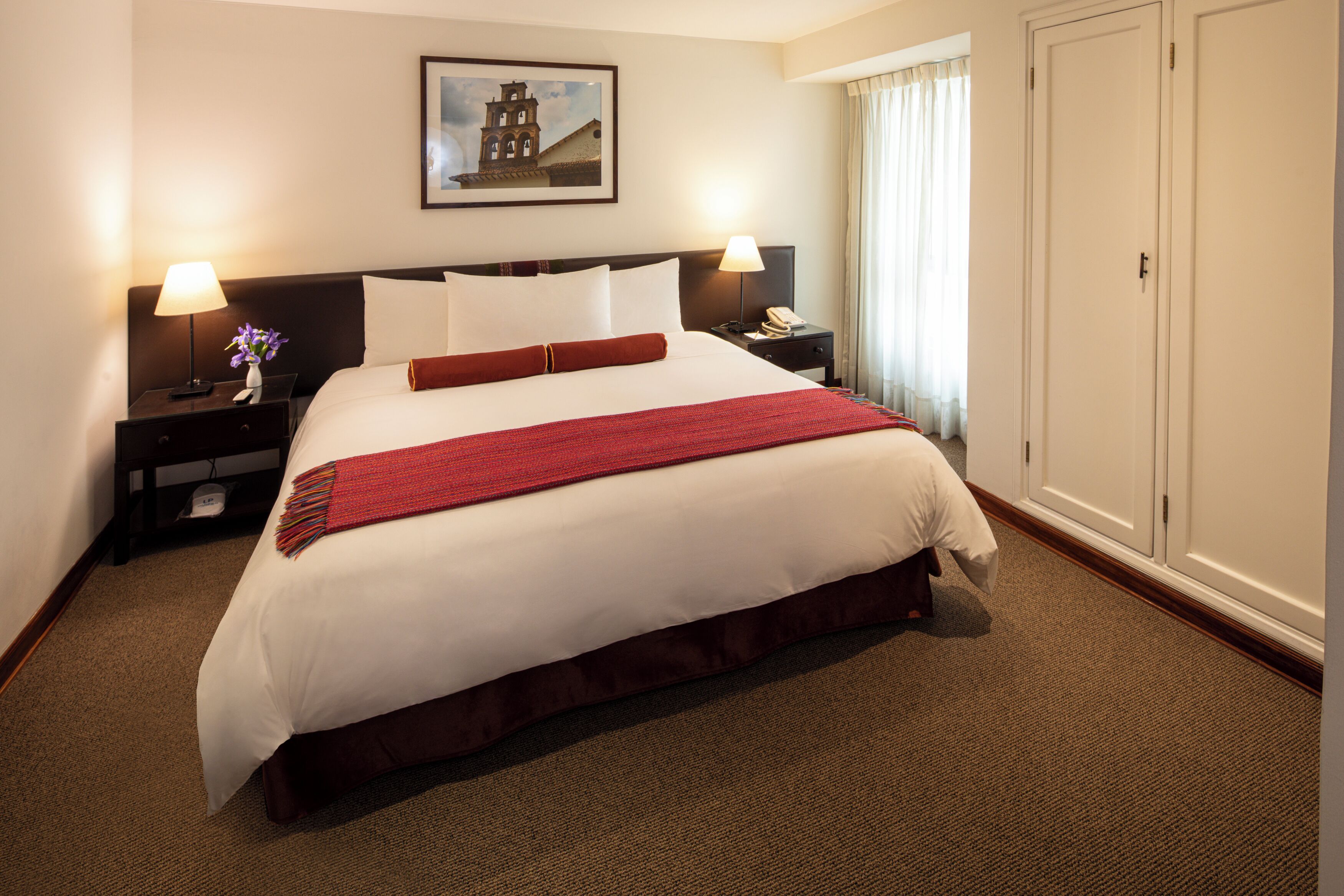 A hotel room with a large bed, two bedside tables with lamps, a nightstand, a framed picture on the wall, and a wardrobe.