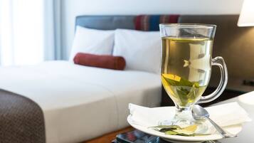 A clear glass mug with a spoon on a white plate, placed on a table in a hotel room with a neatly made bed and a bedside table with a lamp.