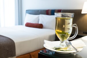 Una taza de vidrio transparente con una cuchara sobre un plato blanco, colocada sobre una mesa en una habitación de hotel con una cama bien hecha y una mesita de noche con una lámpara.
