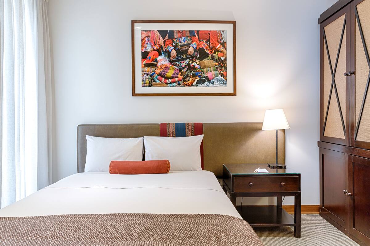 A hotel room with a large bed, a nightstand with a lamp, a framed artwork on the wall, and a wooden wardrobe.