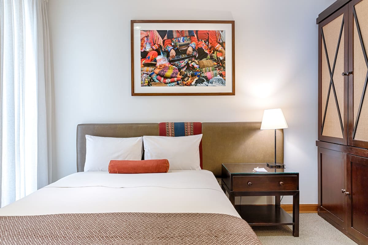 A hotel room with a large bed, a nightstand with a lamp, a framed artwork on the wall, and a wooden wardrobe.