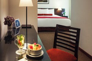 A hotel room with a bed, a desk with a lamp, a chair, and a table with a drink and fruit bowl.