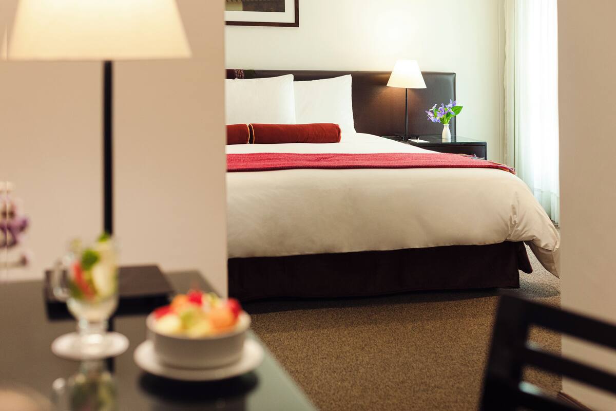 A hotel room with a neatly made bed, a bedside table with a lamp, and a small table with a bowl of fruit.