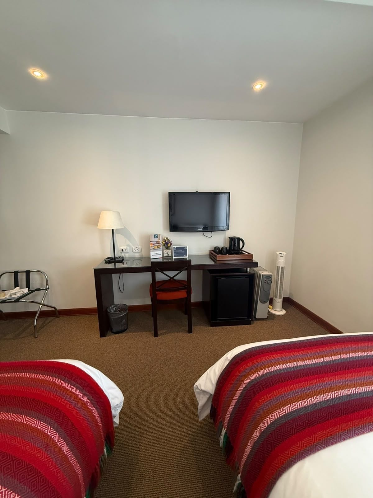 Ein Hotelzimmer mit zwei Betten, einem Schreibtisch mit Computer, einem Fernseher, einem Sessel, einem Mülleimer und einem Heizkörper.