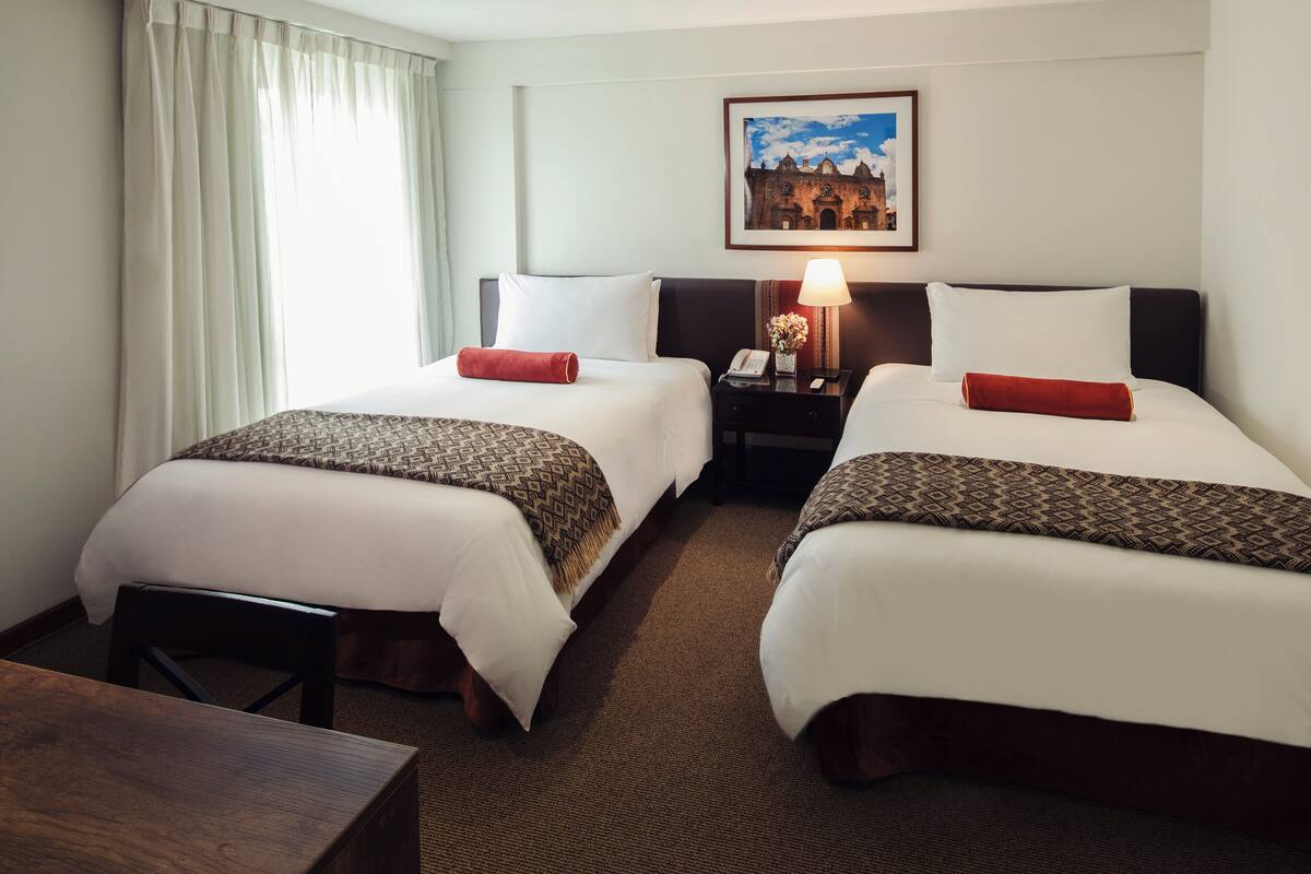 A hotel room with two beds, a nightstand with a lamp, a framed picture on the wall, and a view of a building outside.