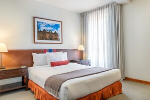 Habitación de hotel con una cama grande, dos mesitas de noche, una lámpara y un cuadro en la pared, además de una ventana con cortinas.