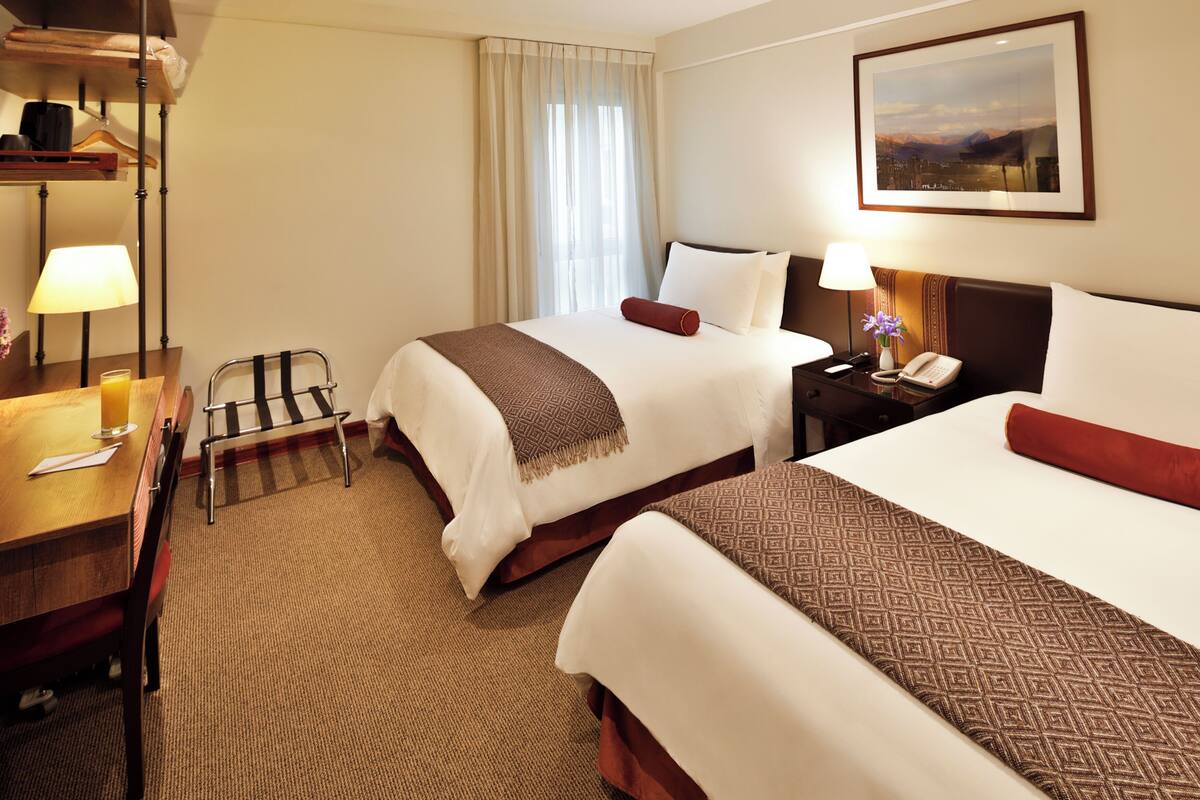 A hotel room with two beds, a desk with a lamp, a chair, a coat rack, and a painting on the wall.