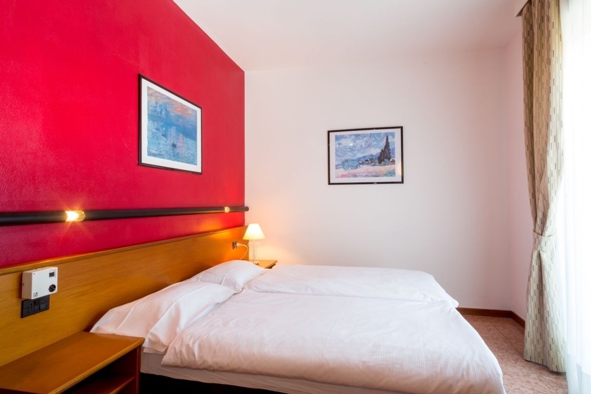 A hotel room with a red accent wall, a bed with white bedding, a nightstand with a lamp, and two framed pictures.