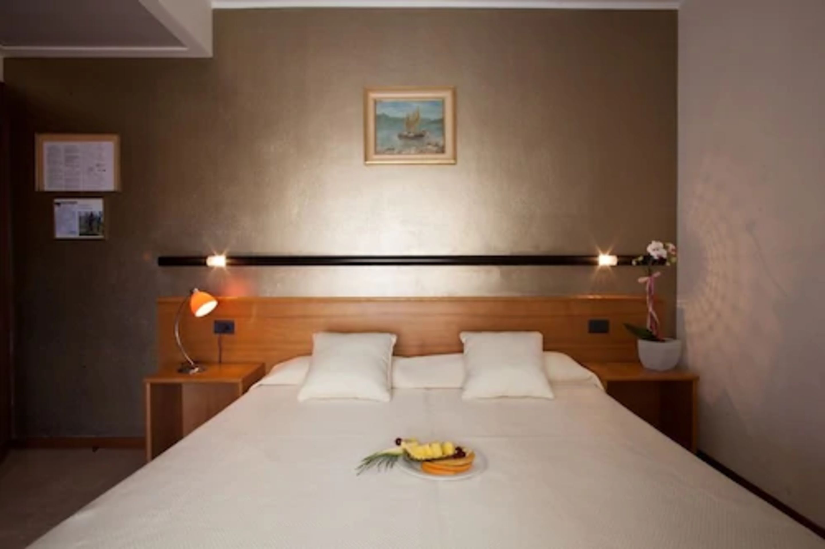 Hotellihuone, jossa on sänky, yöpöydät, lamppu, taulu seinällä ja hedelmäkori sängyllä.