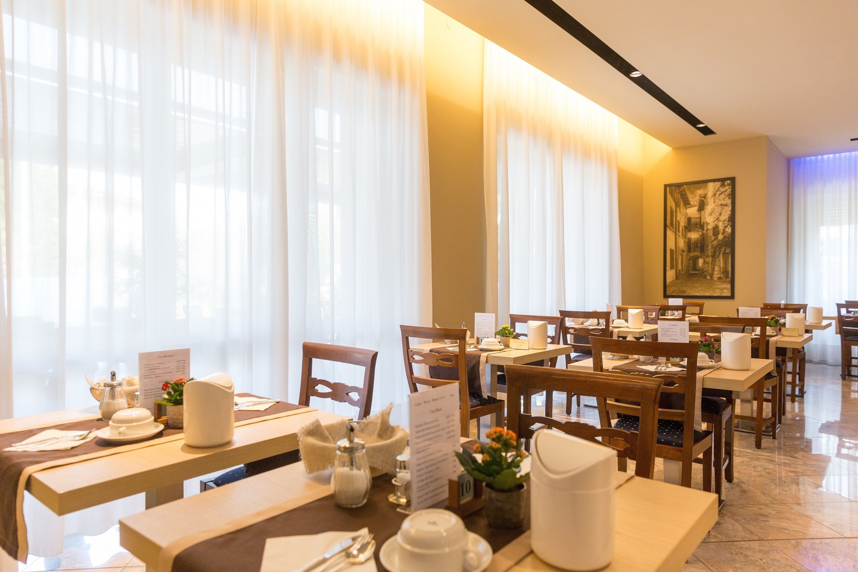 A restaurant with tables set for dining, menus, and a painting on the wall.