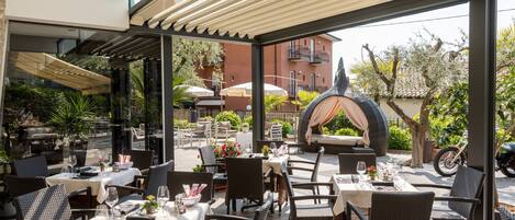 An outdoor dining area with wicker furniture and umbrellas, overlooking a garden and a building with balconies.