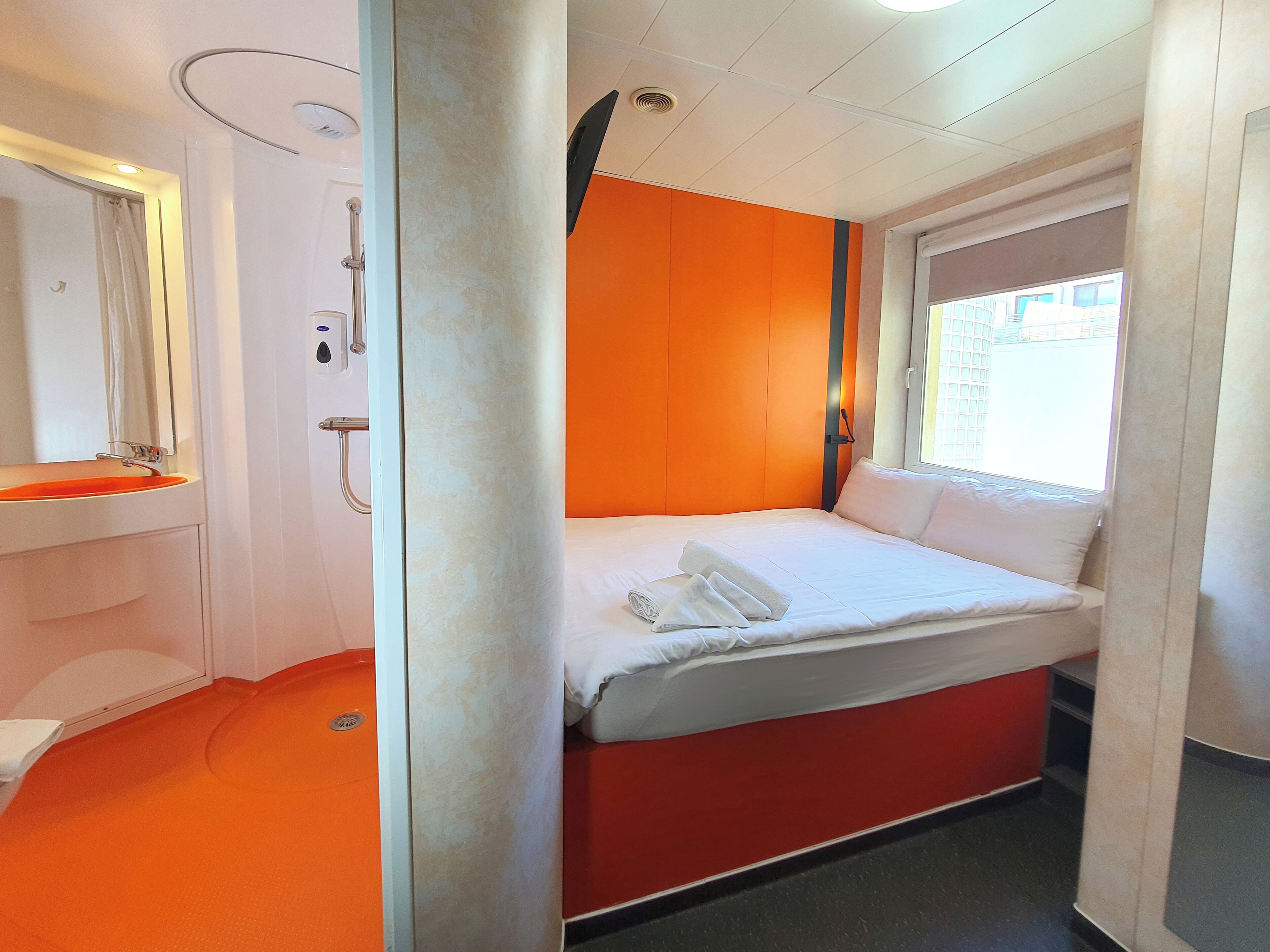 Une chambre d’hôtel avec un lit, une salle de bain dotée d’un lavabo orange et une fenêtre donnant sur des immeubles.