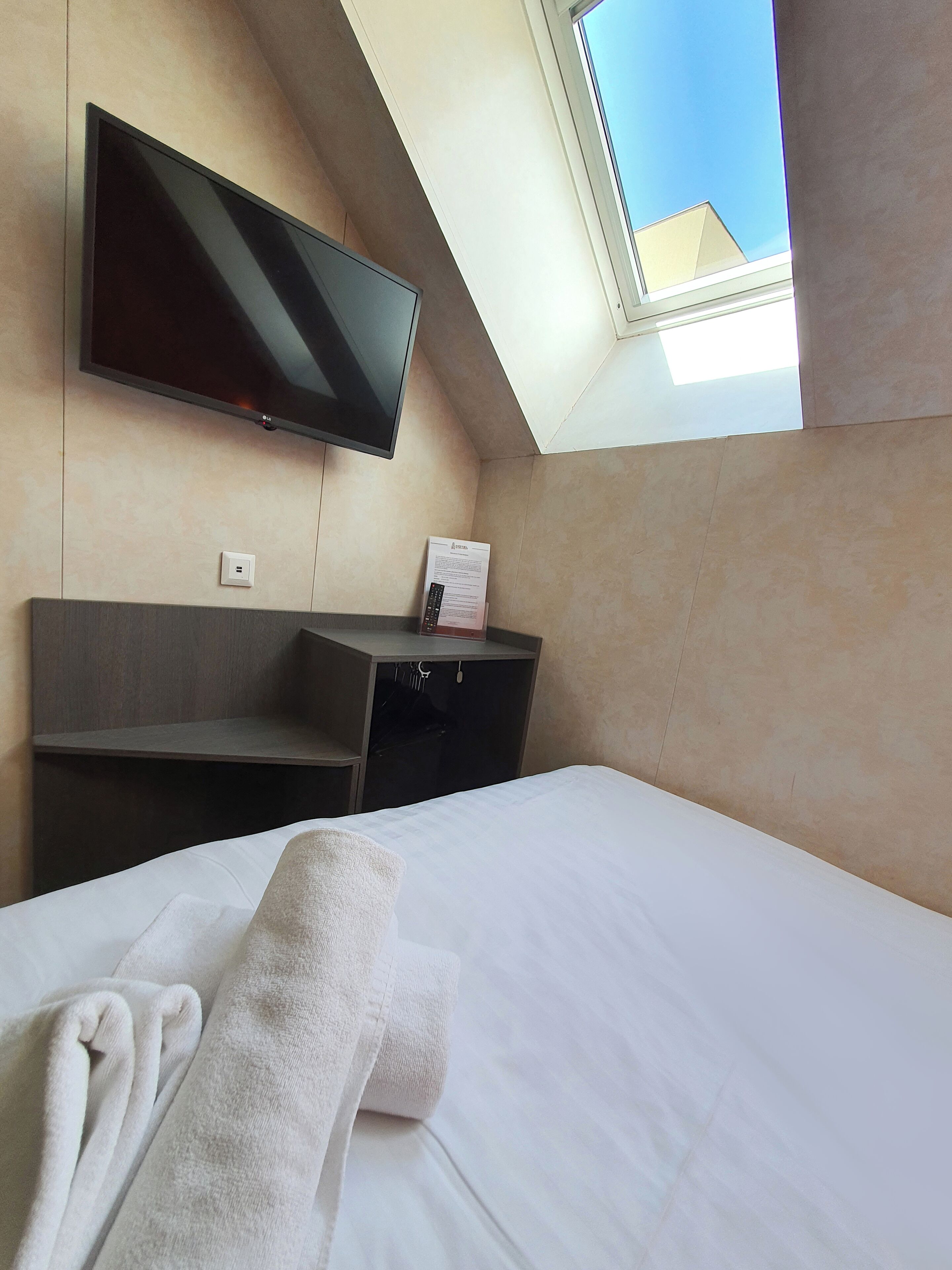 A hotel room with a bed, a flat-screen TV mounted on the wall, a small desk, and a window providing a view of the sky.